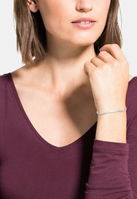 Söljebracelet med en slank, böjd design, med intrikata kedjedetaljer. Bärs på handleden mot en vinröd topp. Slät yta.