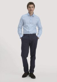 Lichtblauw, langärmelig overhemd met bruine knopen, gecombineerd met donkerblauwe broeken en zwarte schoenen, staand tegen een effen achtergrond.