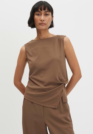 Donna con capelli corti e scuri che indossa un top marrone senza maniche e pantaloni marroni abbinati su uno sfondo chiaro e uniforme.