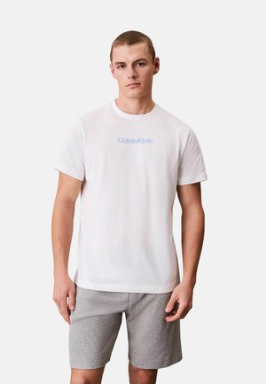 Giovane uomo che indossa una t-shirt bianca Calvin Klein e pantaloncini grigi, in piedi davanti a uno sfondo chiaro uniforme.