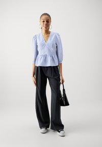 Blå og hvit stripete peplum-bluse med pufferm, kombinert med svarte vide bukser og hvite sneaks, holder en svart veske.