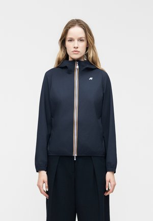 Kobieta o długich włosach, ubrana w granatową kurtkę zapinaną na zamek z małym logo na piersi, stojąca na jednolitym jasnym tle.