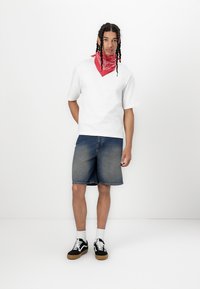 Hvit kortermet skjorte, denimshorts, rød bandana, svarte joggesko med hvite detaljer. Modellen står mot en enkel hvit bakgrunn.