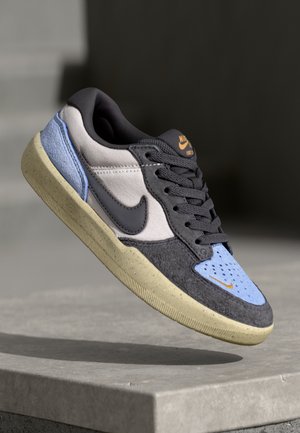 Baskets basses Nike en gris, noir, bleu clair et beige, affichées en suspension au-dessus d'une surface en béton.