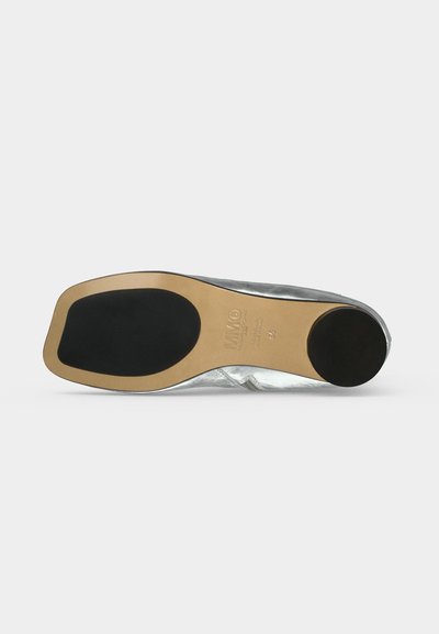 Semelle d'une chaussure argentée avec une base beige et des patins antidérapants noirs au talon et à la plante du pied, présentée sur un fond clair uni.
