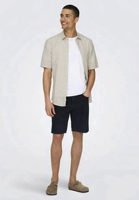Camicetta corta beige chiaro sopra una t-shirt bianca, abbinata a pantaloni corti blu navy e scarpe slip-on beige, si trova di fronte a uno sfondo neutro.