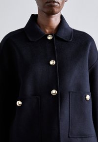 Jūras spēku krāsā, vilnas un sintētisko šķiedru jauktā mēteli ar apkakles izgriezumu, ar zelta pogām un priekšējām kabatām. Audumam ir mīksta, teksturēta virsma.