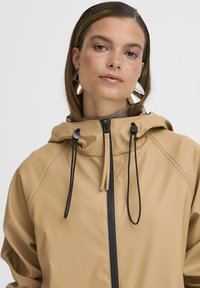 Veste imperméable beige avec capuche, dotée d'une fermeture éclair à l'avant, de cordons de serrage noirs et d'une texture lisse. Accentuée par un col haut.
