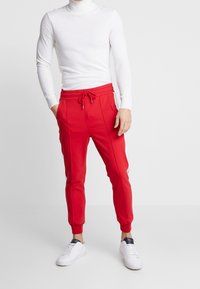 Mann, der rote Jogginghosen mit weißen Seitenstreifen, einen weißen figurbetonten Rollkragenpullover und weiße Sneakers trägt und vor einem schlichten Hintergrund steht.
