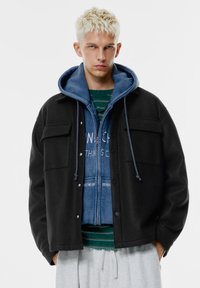 Černá bunda z vlněné směsi se dvěma n�áprsními kapsami, nošená přes modrou mikinu na zip. Model má pod ní pruhované zelené tričko. Jsou vidět šedé tepláky.