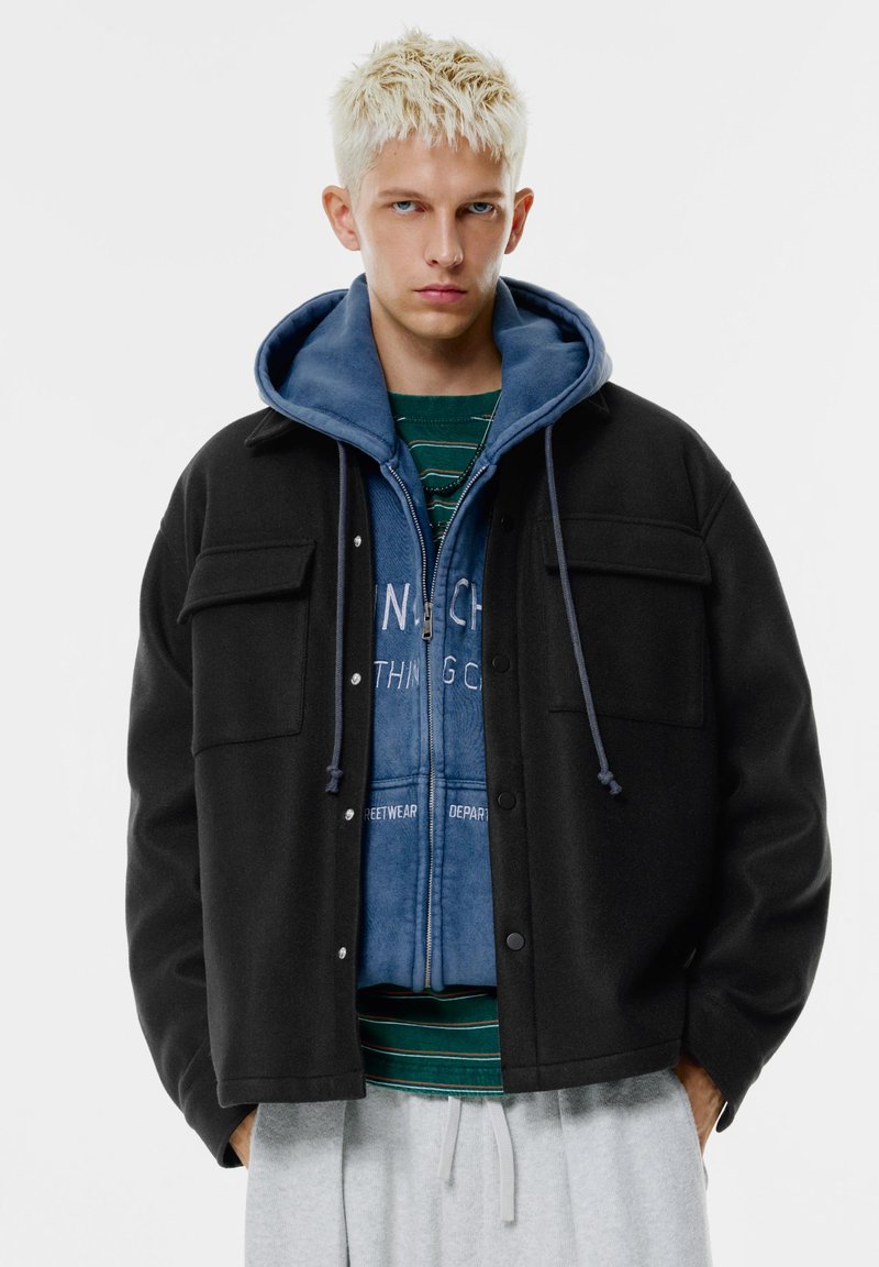 Černá bunda z vlněné směsi se dvěma náprsními kapsami, nošená přes modrou mikinu na zip. Model má pod ní pruhované zelené tričko. Jsou vidět šedé tepláky.