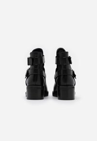 Bottines noires en cuir avec un talon épais, dotées de deux sangles réglables et d'une finition lisse et brillante. Le design est minimaliste.