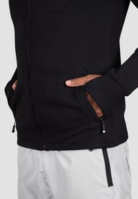 Schwarze Zip-Jacke mit glatter Textur, gerippten Bündchen und seitlichen Taschen. Kombiniert mit hellen Hosen für einen Kontrast.