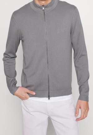 Muž nosící světle šedý svetr na zip přes bílou košili, s jednou rukou v kapse a v bílých kalhotách.