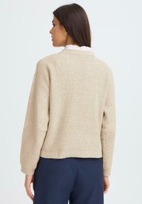 Beżowy sweter z dzianiny o prążkowanej fakturze, krótki i z szerokimi rękawami. Posiada biały, falbankowy kołnierz wystający spod spodu.