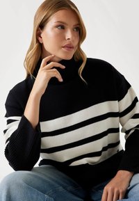 Maglione a collo alto a righe bianco e nere con polsini a costine, caratterizzato da una vestibilità comoda e una texture lavorata a maglia. Abbinato a jeans in denim azzurro chiaro.