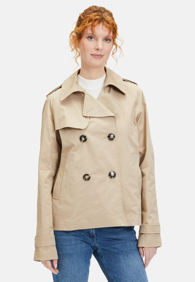 Beige double-breasted trench coat with a wide collar, large black buttons, and side pockets; made from smooth, lightweight fabric.