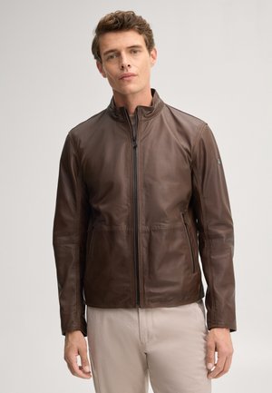 Junger Mann trägt eine braune Lederjacke mit Reißverschluss und Seitentaschen sowie beige Hosen, steht vor einem einfarbigen hellen Hintergrund.