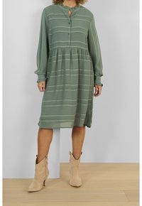 Abito verde a righe con colletto, chiusura con bottoni e polsini elasticizzati. Indossato con stivali da cowgirl beige, presenta una gonna leggermente svasata.