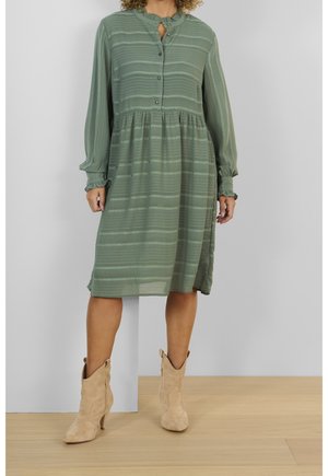 Robe à rayures vertes avec un col, fermeture à boutons et manches élastiques. Portée avec des bottes de cowgirl beiges, présentant une jupe légèrement évasée.