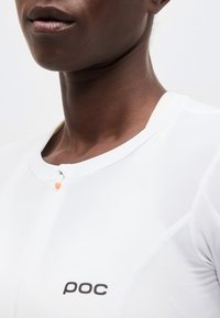 Κοντινό πλάνο ενός ατόμου που φοράει λευκό αθλητικό τοπ με το λογότυπο της POC και μικρό πορτοκαλί φερμουάρ στο γιακά.