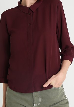 Kvinde iført en langærmet, knappet bordeauxbluse med opkrøllede ærmekanter og grønne bukser, med hånden stukket i lommen.