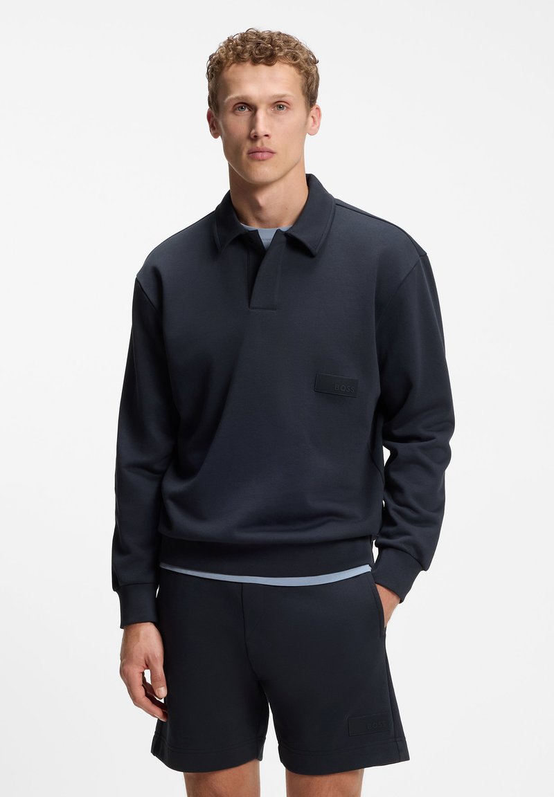 Jeune homme aux cheveux bouclés portant un polo à manches longues bleu marine foncé et un short assorti sur un fond uni.