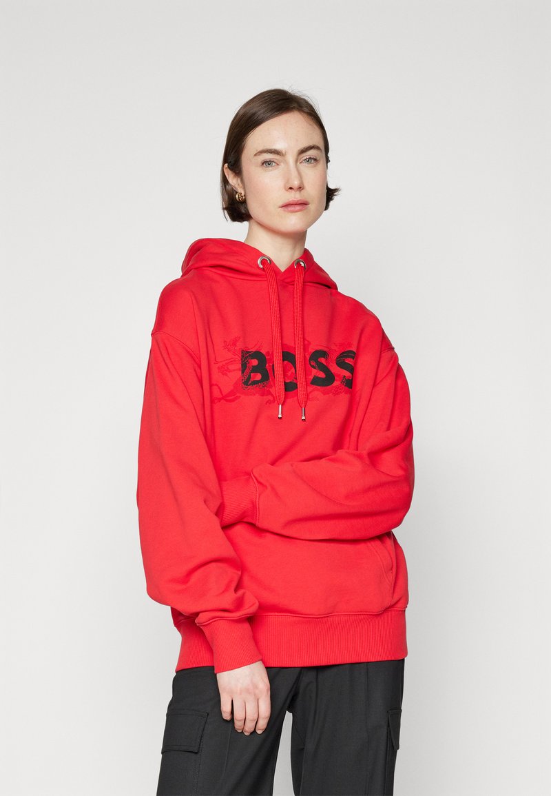 Sweat-shirt en coton rouge avec un grand logo noir "BOSS", capuche avec cordon de serrage et poignets côtelés. Le modèle porte un pantalon cargo noir.