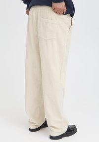 Beżowe sztruksowe spodnie o teksturowanej powierzchni, luźnym kroju, elastycznej talii i tylną kieszenią. Połączone z czarnymi butami.