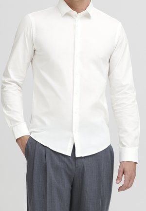 Homme portant une chemise blanche unie à manches longues et un pantalon gris à fines rayures, debout avec une main dans la poche.