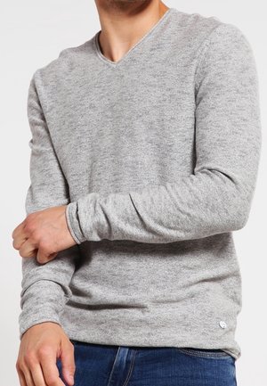 Mann trägt einen hellgrauen Langarm-V-Ausschnittpullover und zieht den linken Ärmel hoch, bekleidet mit blauen Jeans vor einem einfarbigen Hintergrund.