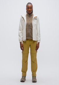 Λευκό αδιάβροχο μπουφάν με κουκούλα πάνω από καφέ φλις με φερμουάρ, συνδυασμένο με ταμπά παντελόνια cargo και γκρι παπούτσια πεζοπορίας.