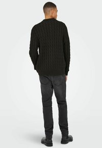 Donkergroene gebreide trui met een kabelpatroon, gedragen met zwarte jeans en zwarte schoenen, gezien van achteren tegen een lichte achtergrond.