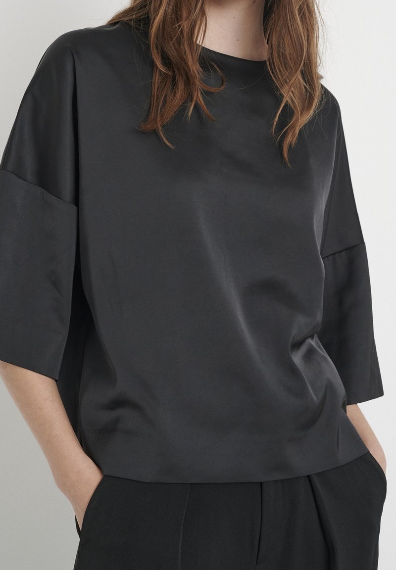 Femme portant une chemise noire ample à manches courtes et un pantalon noir assorti, les mains dans les poches, vue du cou jusqu’aux hanches.