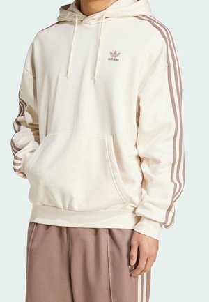 Person trägt einen cremefarbenen Adidas-Hoodie mit pinken Streifen an den Ärmeln und passende pinke Adidas-Trainingshose mit weißen Streifen.