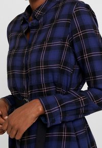 Marinblå rutig skjorta med rosa och svarta mönster, med en knappad krage, två bröstfickor och bältesdetaljer vid midjan.