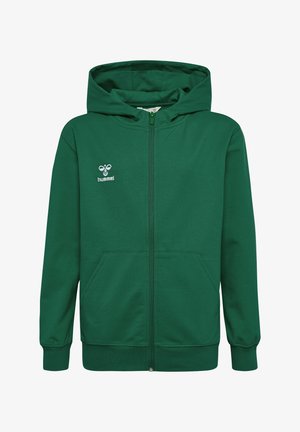 To børn står iført grønne hættetrøjer, sorte skjorter, sorte bukser og sorte sneakers mod en ensfarvet baggrund.