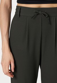 Pantalons en tissu côtelé vert foncé avec une taille à cordon, présentant des plis et des poches latérales pour un look sophistiqué. La texture est lisse et structurée.