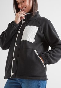 Black fleece jacket with white accents, a chest pocket, snap buttons, high collar, and elastic cuffs. Soft texture with a relaxed fit.