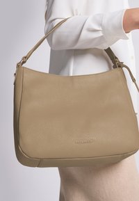 Beige Leder Schultertasche mit glatter Textur, runder Form und verstellbarem Tragegurt, ausgestattet mit einem unauffälligen Logo am Boden.