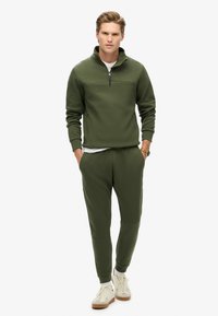 Sudadera de forro polar verde oliva con media cremallera, combinada con pantalones tapered a juego. Tejido ligero, diseño minimalista, acentuado con zapatillas blancas.