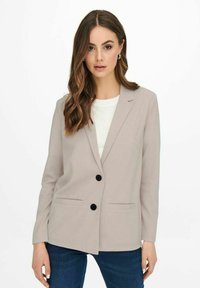 Světle béžový blazer s dvěma černými knoflíky, s klopami, postranními kapsami a strukturovaným střihem, nošený přes bílý top a modré džíny.