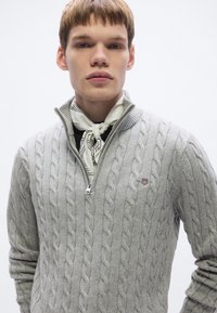 Ανοιχτό γκρι πουλόβερ με πλέξη καλώδιο και φερμουάρ στον γιακά, με ένα μικρό κεντημένο λογότυπο. Ένα λευκό κασκόλ με μαύρο σχέδιο δένεται στον λαιμό.