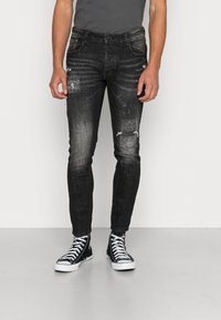 Jeans de mezclilla negros con corte ajustado, con detalles desgastados y salpicaduras de pintura. Llevados con zapatillas altas negras.