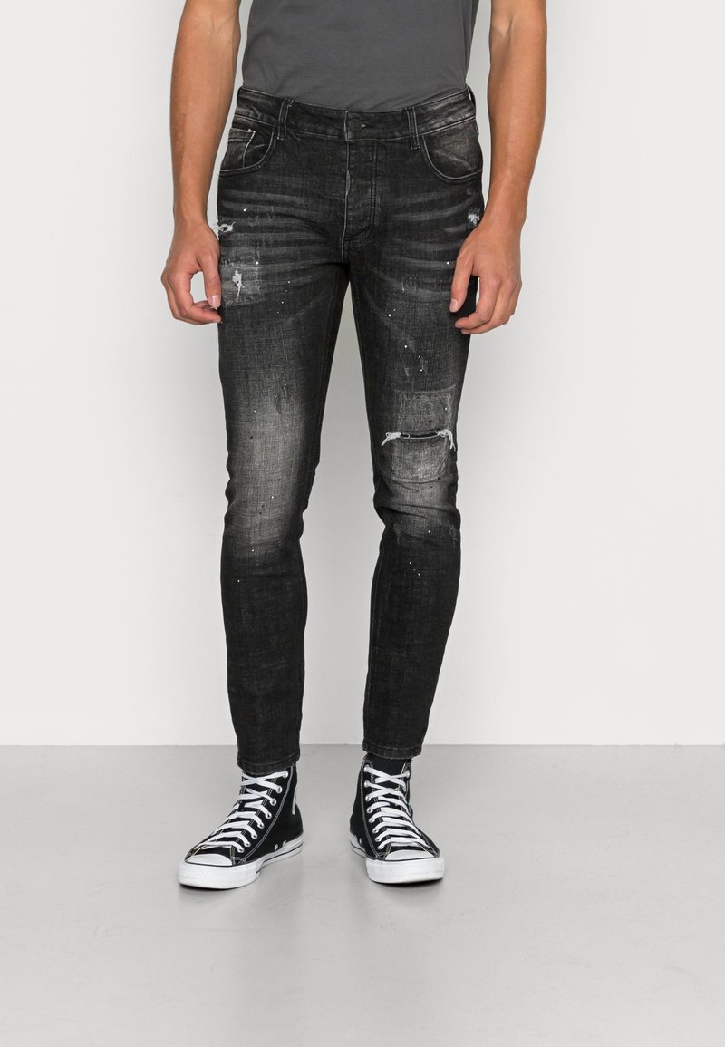 Jeans de mezclilla negros con corte ajustado, con detalles desgastados y salpicaduras de pintura. Llevados con zapatillas altas negras.