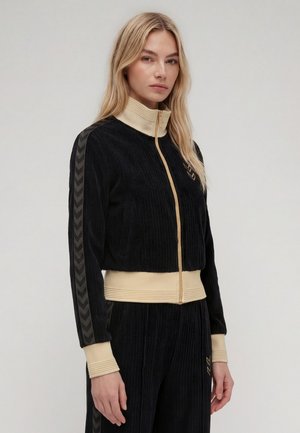 Femme blonde portant un survêtement en velours noir avec col, poignets et ceinture beige, debout devant un fond clair uni.