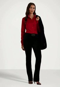 Γυναίκα που φοράει ένα κόκκινο πουκάμισο με κουμπιά, μαύρο παντελόνι με διακοσμητική ζώνη, μαύρες ψηλές γόβες και κρατά μια μαύρη ζακέτα πάνω στον ώμο της.