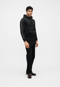 Homme debout portant un sweat à capuche noir avec des bandes latérales, un pantalon noir et des chaussures habillées marron, sur un fond clair uni.