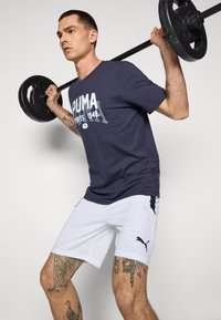 Puma donkerblauwe t-shirt met witte logo en 1948 afdruk; witte shorts met zwarte accenten en logo; persoon die een halter heft.