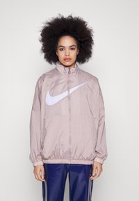 Ljusrosa Nike vindjacka med hög krage, långa ärmar, elastiska muddarna och en stor vit swoosh-logotyp på framsidan.